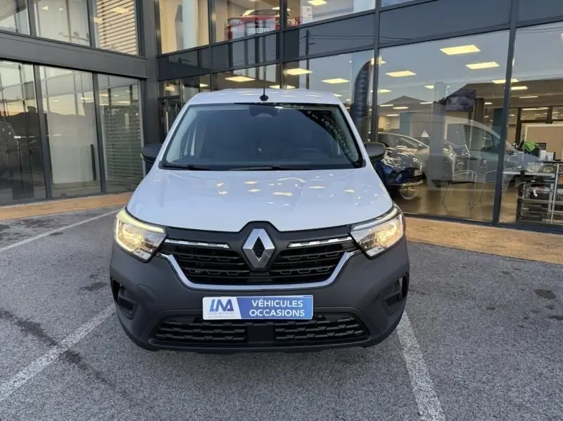 Vue avant d'un Renault Kangoo blanc 2022 avec phares allumés, devant une vitrine de concession.