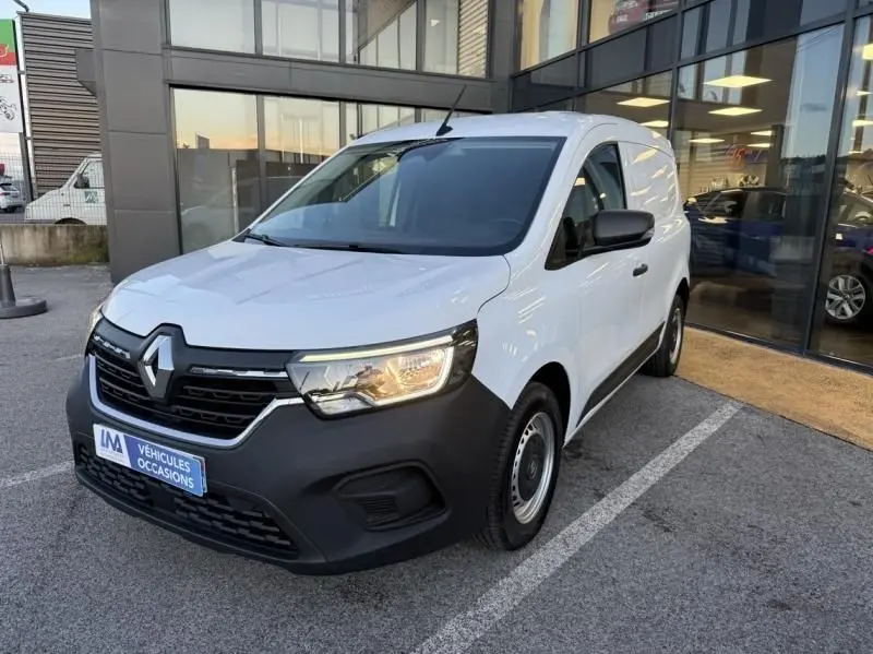 Renault Kangoo Van DCI blanc vu en 3/4 avant droit, avec pare-chocs noir mat et phares allumés, stationné devant un bâtiment.