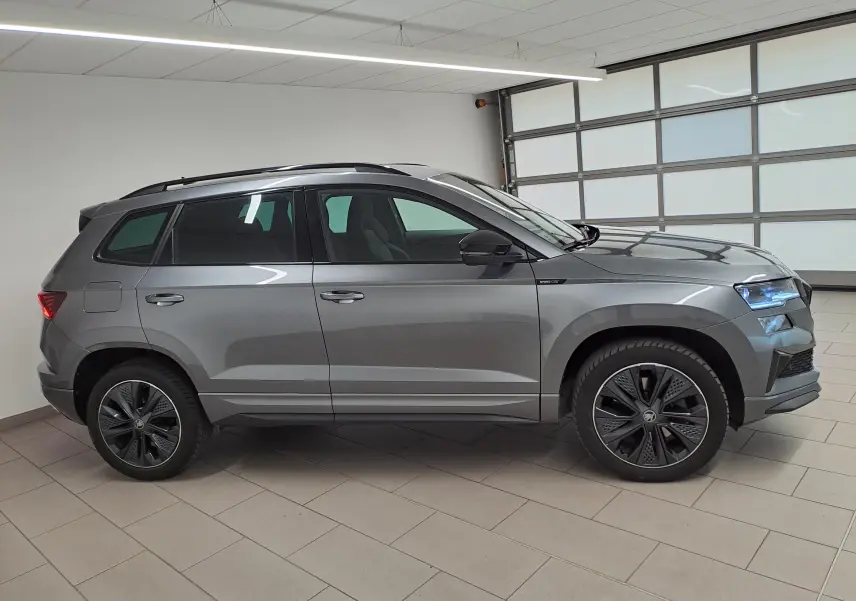Vue de profil côté gauche d'un SKODA KAROQ gris graphite métallisé, avec jantes noires et toit ouvrant, en intérieur showroom.