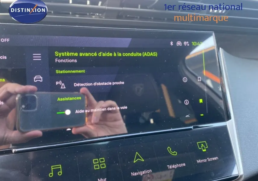 Écran tactile intérieur du Peugeot 308 Hybrid 145ch GT montrant les aides à la conduite, reflet de mains et smartphone visible.