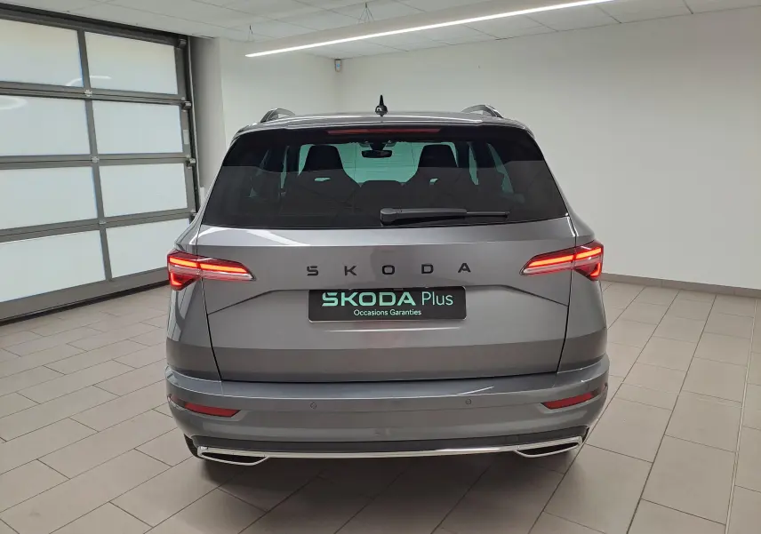Vue arrière d'un SKODA KAROQ gris graphite 2025 avec feux allumés et hayon électrique en intérieur showroom.
