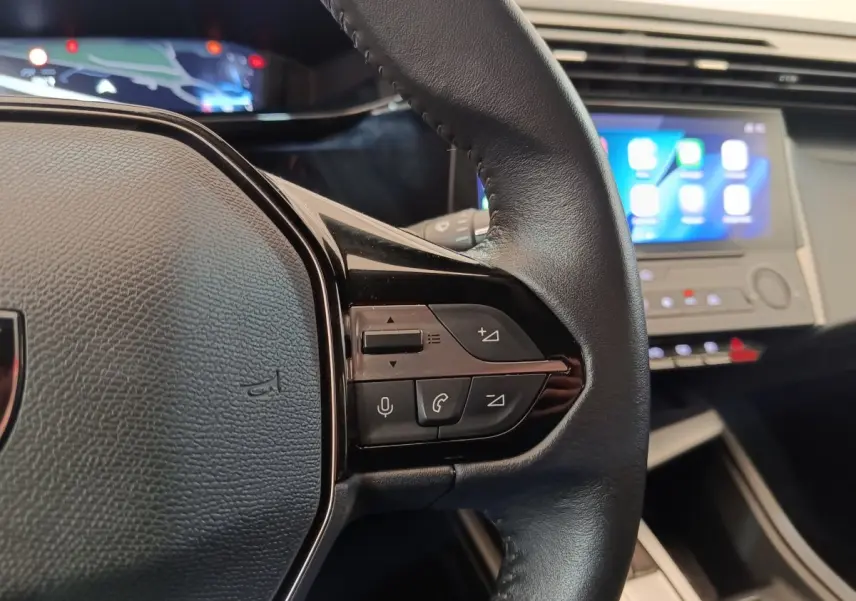 Gros plan sur le volant cuir noir côté droit de la Peugeot 308 2023 avec commandes vocales et écran tactile en arrière-plan.