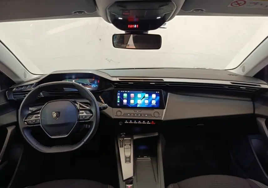 Vue intérieure avant de la Peugeot 308 2023, tableau de bord noir avec écran tactile central lumineux et volant cuir multifonctions.