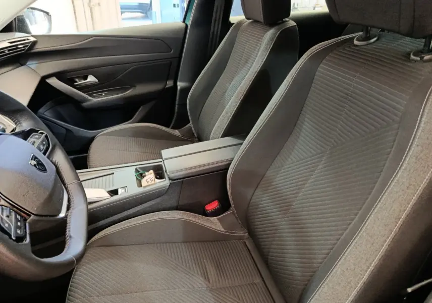 Vue intérieure côté conducteur de la Peugeot 308 2023, sièges tissu gris et console centrale avec accoudoir.