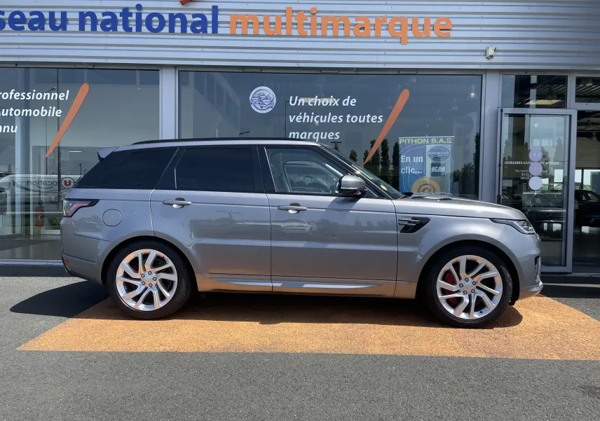 Vue latérale droite d'un Range Rover Sport gris Eiger avec jantes alu 21 pouces et vitres arrière surteintées, stationné devant un showroom.
