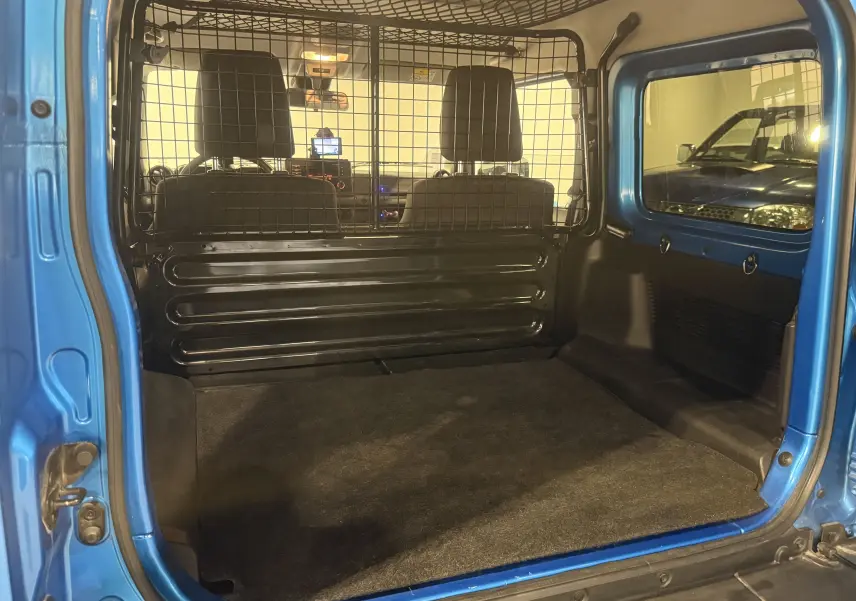 Vue arrière intérieure du Suzuki Jimny bleu 2021, montrant le coffre avec séparation grillagée et sièges avant.