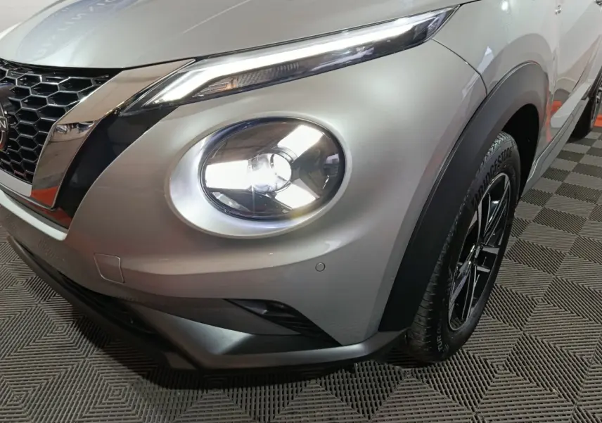 Vue 3/4 avant droit du Nissan Juke gris foncé avec phare rond allumé et jante noire à motifs distinctifs.