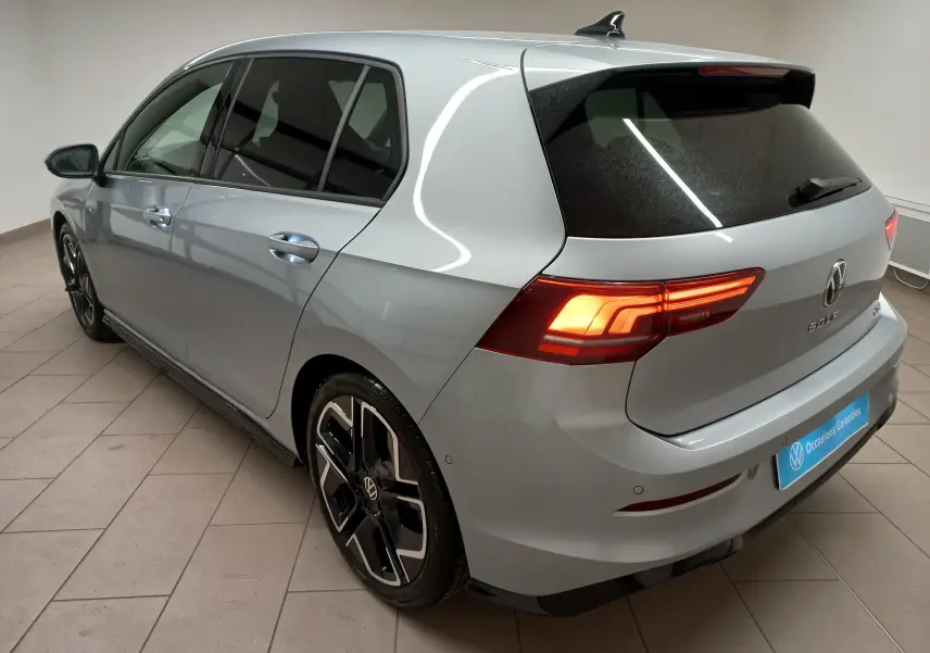 Vue 3/4 arrière gauche d'une Volkswagen Golf 1.5 eTSI R-Line argent dolomite avec feux allumés et jantes spécifiques.