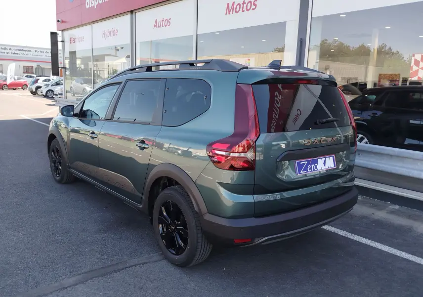 Vue 3/4 arrière droite d'un Dacia Jogger 2026 vert cèdre métallisé avec jantes noires et barres de toit, stationné devant un showroom.