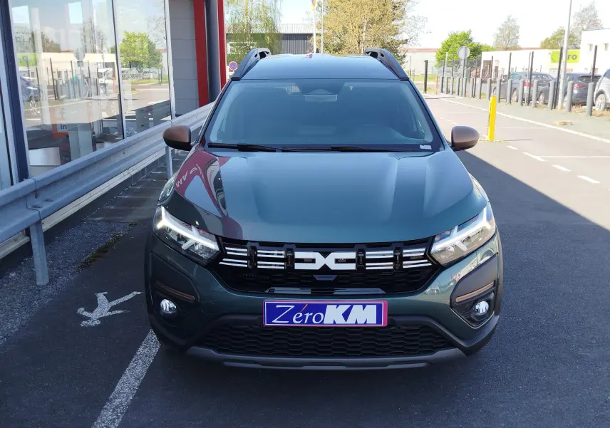 Vue avant d'un Dacia Jogger 2026 vert cèdre métal avec calandre noire et phares LED allumés, stationné devant un bâtiment.