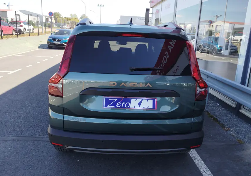 Vue arrière d'un Dacia Jogger vert cèdre métal 2026 avec logo et plaque "Zero KM" sur un parking extérieur.