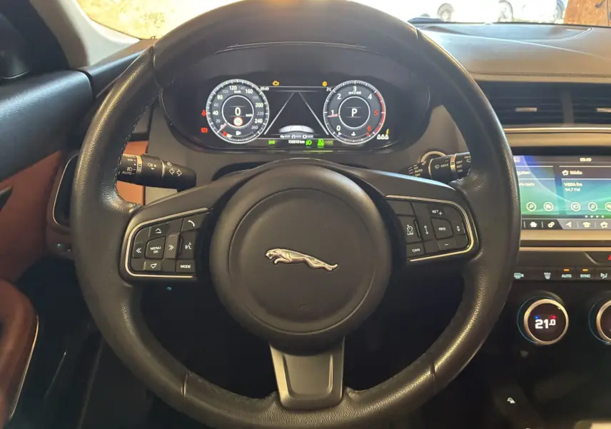 Vue intérieure centrée sur le volant noir avec logo Jaguar, tableau de bord numérique et écran multimédia visible à droite.