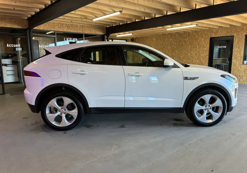 Jaguar E-PACE blanc vue de profil côté gauche, SUV compact avec jantes alliage et toit plongeant dans un showroom.