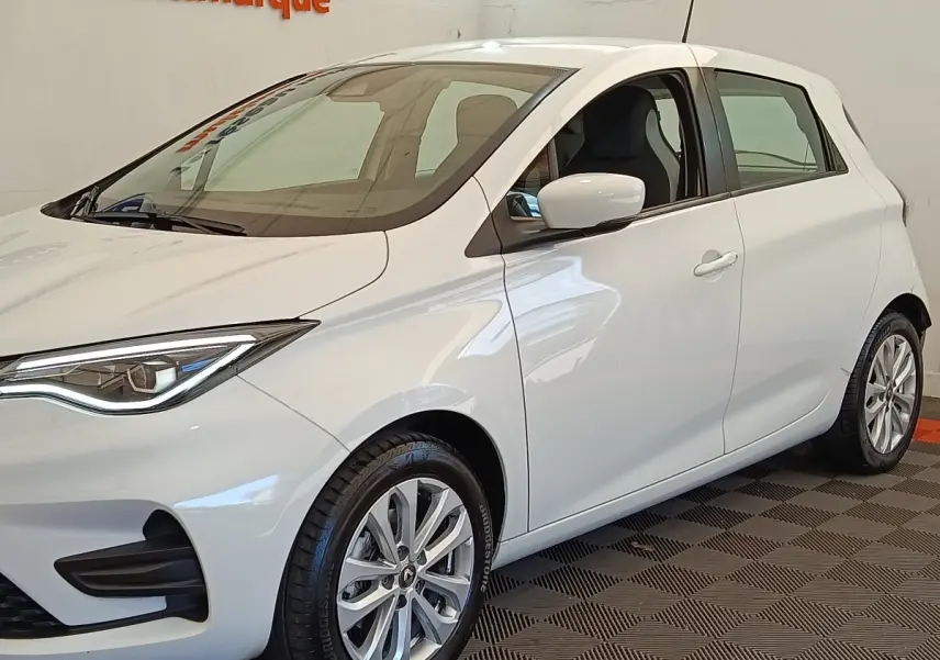 Renault ZOE blanc vue 3/4 avant droit, phares LED et jantes alliage visibles en intérieur showroom.