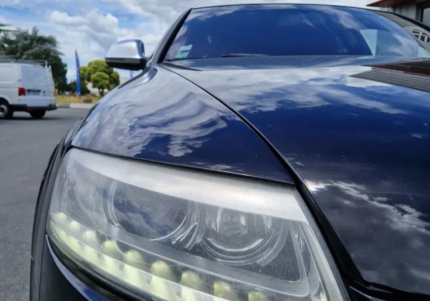 Gros plan sur le phare avant gauche noir brillant d'une Audi RS6 Avant 2009, reflétant le ciel nuageux.