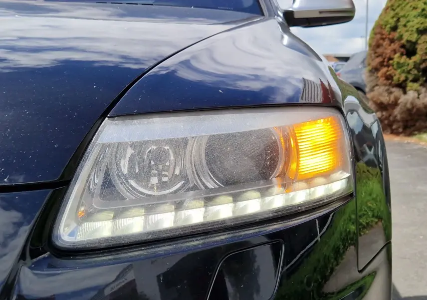 Gros plan sur le phare avant droit allumé d'une Audi RS6 Avant noire de 2009, reflétant l'environnement extérieur.