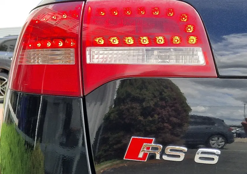 Gros plan sur le feu arrière droit rouge et le logo RS6 d'une Audi RS6 Avant noire, reflet d'un paysage urbain visible.