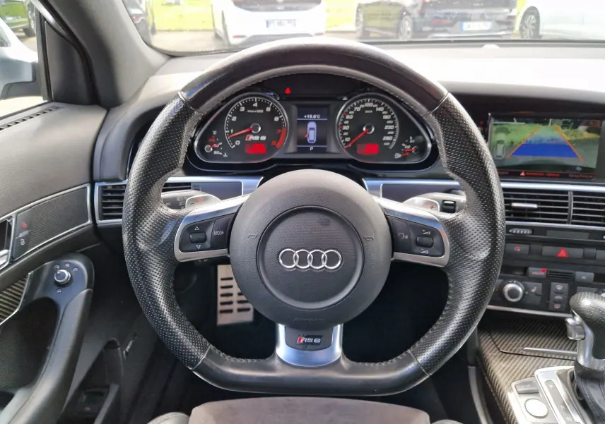 Vue intérieure centrée sur le volant en cuir perforé noir d'une Audi RS6 Avant 2009 avec tableau de bord et écran caméra de recul.