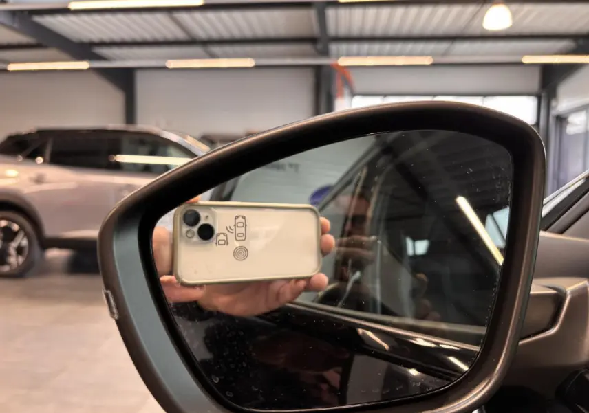 Rétroviseur extérieur droit noir brillant du Peugeot 2008 blanc, reflétant partiellement l'intérieur et un smartphone.