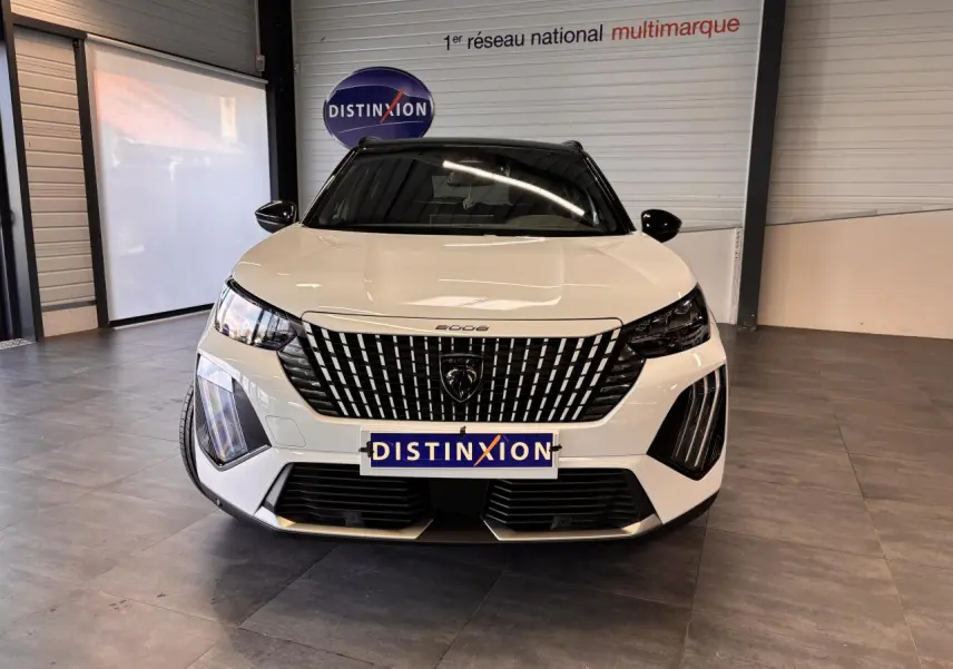 Vue frontale d'un Peugeot 2008 blanc avec calandre noire à motifs verticaux et phares LED distinctifs en intérieur showroom.