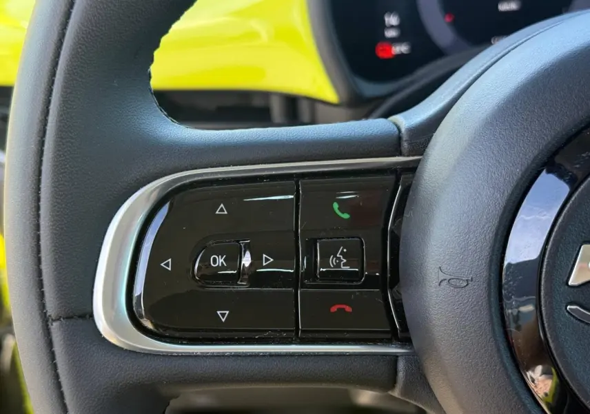 Gros plan sur les commandes au volant à gauche d'une Fiat 500 e jaune, avec tableau de bord numérique en arrière-plan.