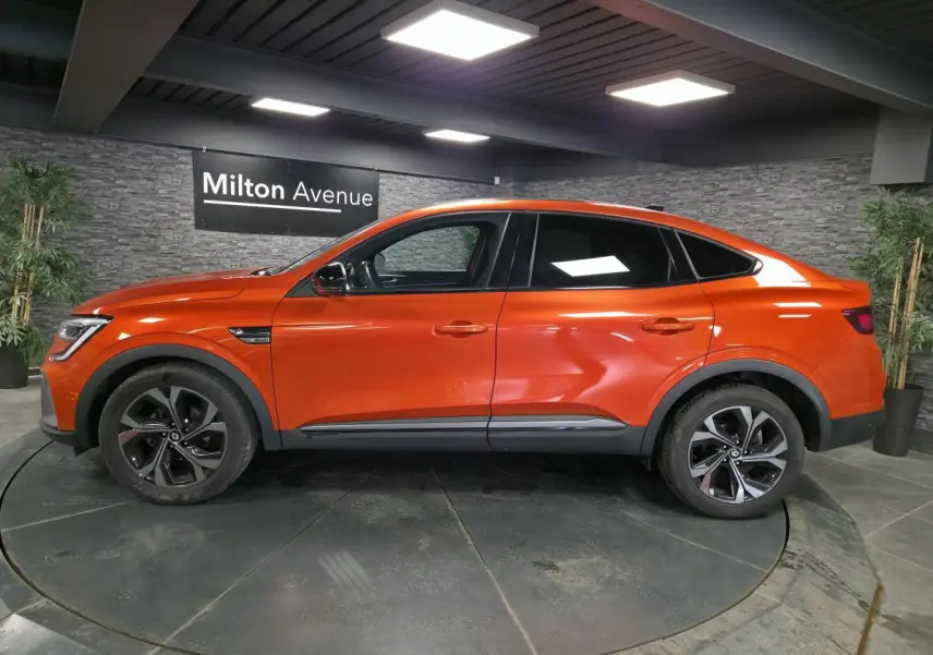 Profil droit du Renault Arkana 2024 orange Valencia avec jantes 18 pouces et toit ouvrant électrique visible en intérieur showroom.