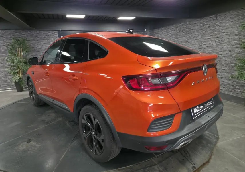 Vue 3/4 arrière droite d'un Renault Arkana orange Valencia avec jantes noires et toit ouvrant en intérieur showroom.
