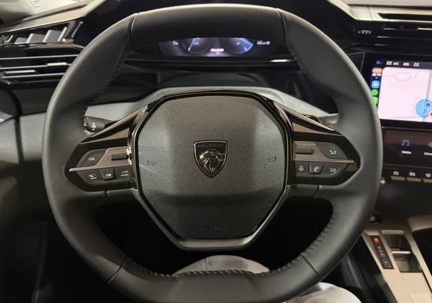 Vue rapprochée du volant cuir noir de la Peugeot 308 2025 avec commandes multifonctions et écran tactile en arrière-plan.