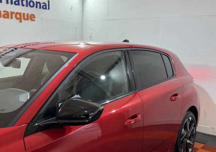 Vue latérale gauche d’une Peugeot 308 rouge 2025 avec rétroviseur noir et vitres teintées.