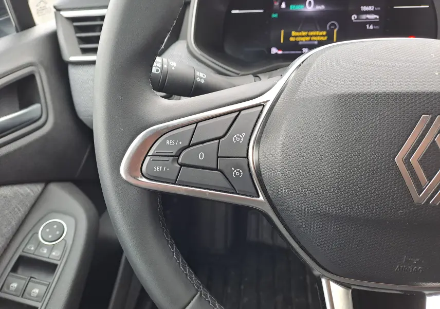 Gros plan sur le volant multifonction noir de la Renault Clio V 2025, avec commandes de régulateur et tableau de bord numérique en arrière-plan.