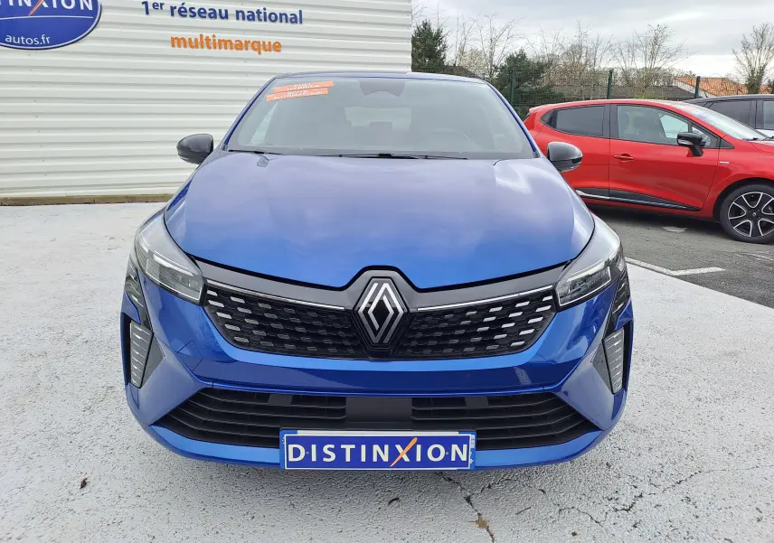 Vue frontale d'une Renault Clio V bleu Iron Metal avec calandre noire et logo Renault proéminent, stationnée en extérieur.