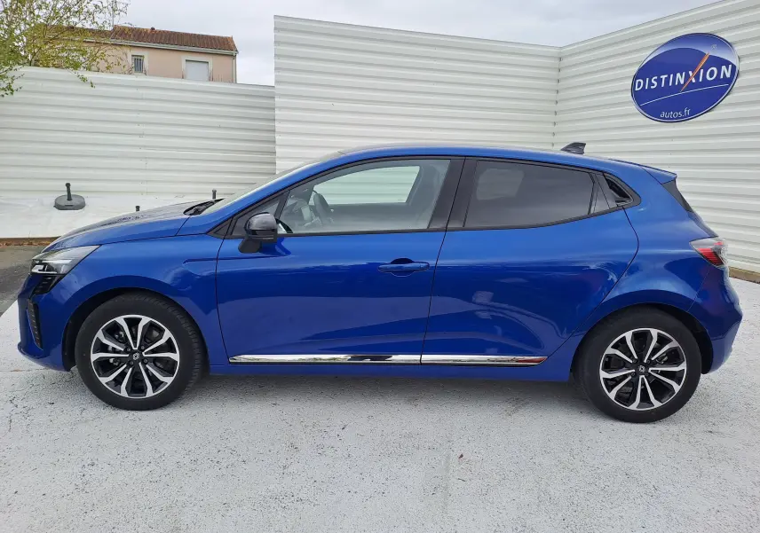Profil droit de la Renault Clio V hybride bleu Iron Metal 2025 avec jantes alliage et détails chromés sur bas de caisse.