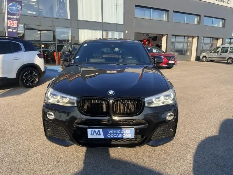 BMW X4 30DA X DRIVE noir métal vu de face, avec calandre noire et phares LED allumés, sur parking extérieur.