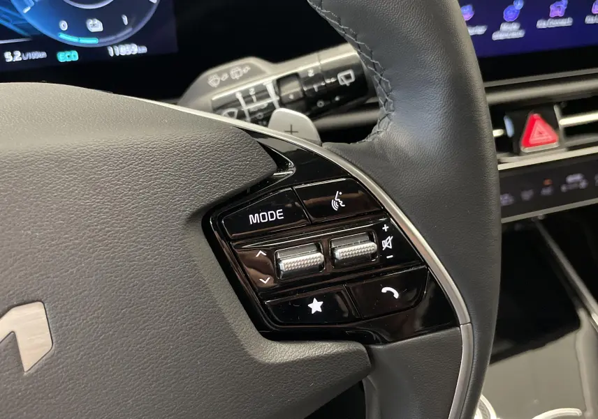 Gros plan sur le volant cuir noir du KIA NIRO blanc 2025, avec commandes multifonctions et palettes au volant visibles.