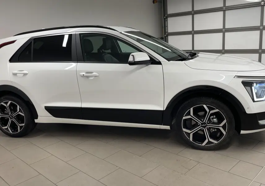KIA NIRO hybride blanc vu de profil côté gauche, avec jantes noires et phares LED allumés dans un garage.