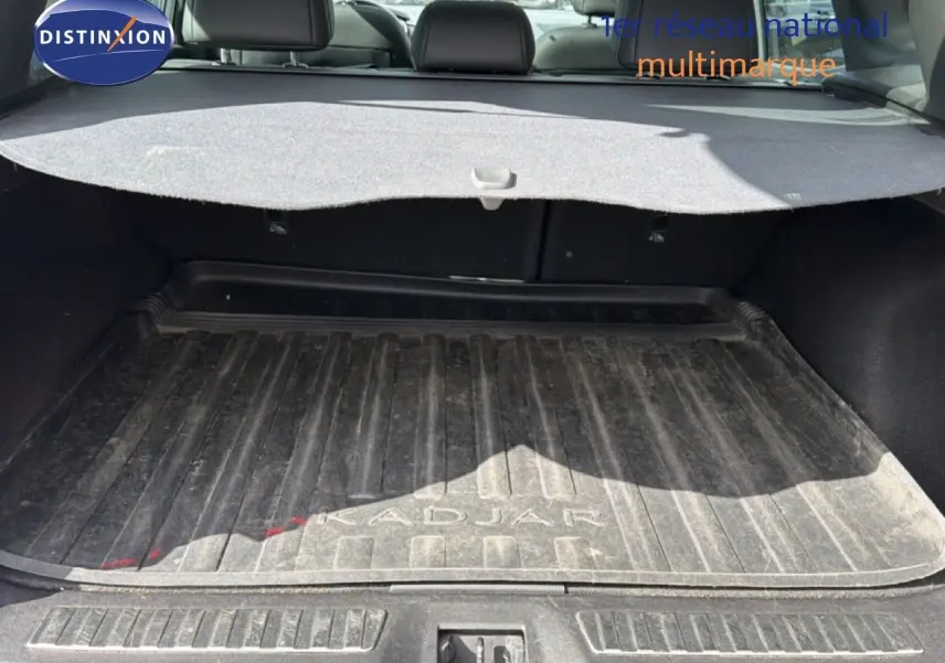 Coffre ouvert vu de l'arrière d'un Renault Kadjar gris Highland avec tapis de coffre marqué Kadjar et cache-bagages déployé.