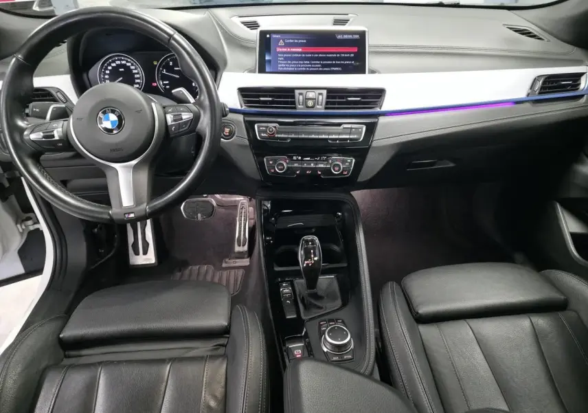 Vue intérieure avant du BMW X2 sDrive 18d M Sport 2023, avec sellerie cuir noire et éclairage d'ambiance violet sur la planche de bord.