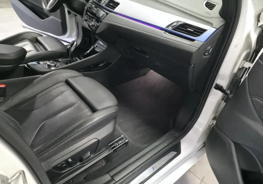 Vue intérieure côté passager avant d'une BMW X2 blanche, mettant en valeur la sellerie cuir noire et l'éclairage d'ambiance bleu.