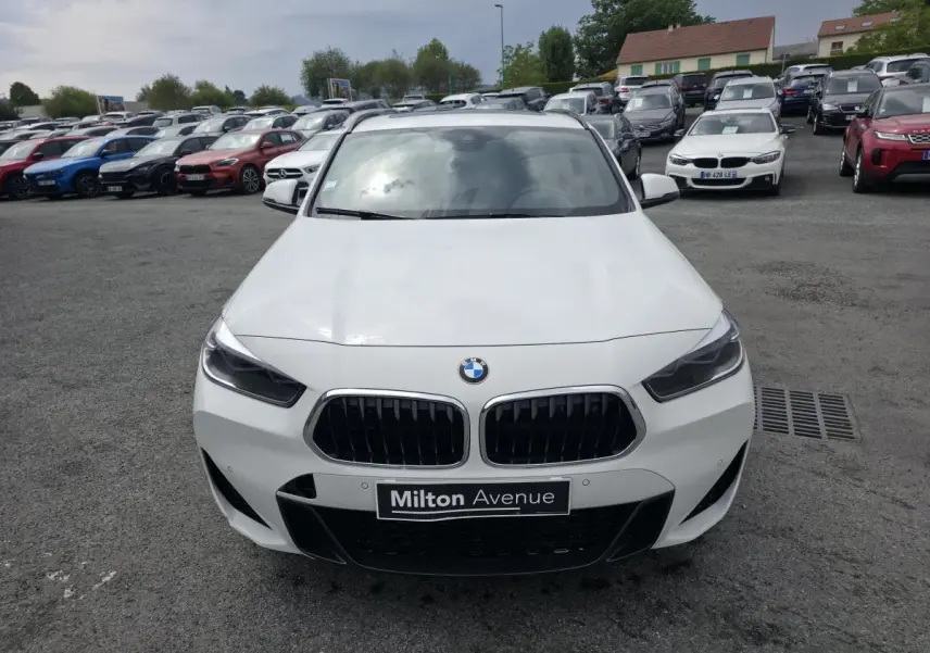 Vue avant d'une BMW X2 sDrive 18d blanche avec calandre noire et plaque Milton Avenue visible en concession.
