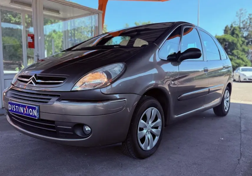 Vue 3/4 avant droit d'une Citroën Xsara Picasso beige métallisé garée sous un abri avec phares allumés.