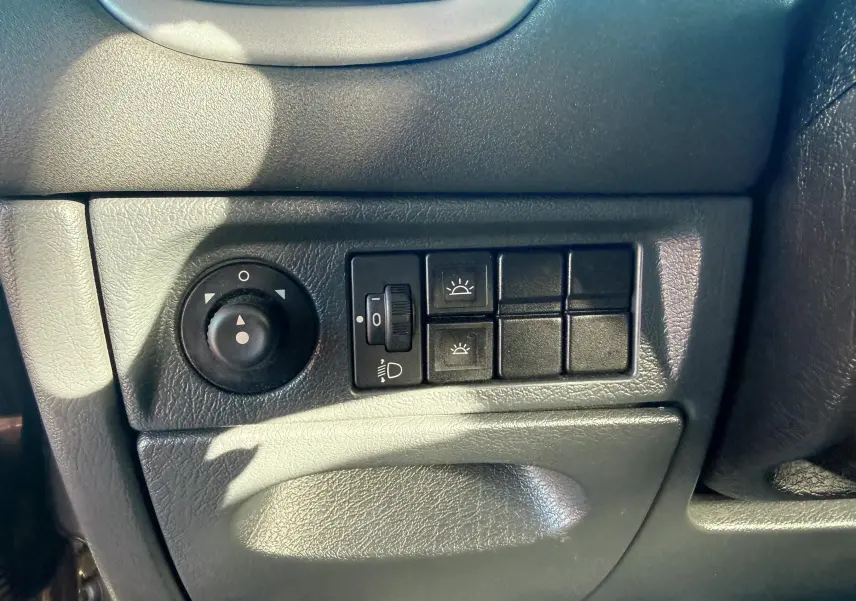 Gros plan sur les commandes d’éclairage et réglage des rétroviseurs du tableau de bord noir d’une Citroën Xsara Picasso beige.