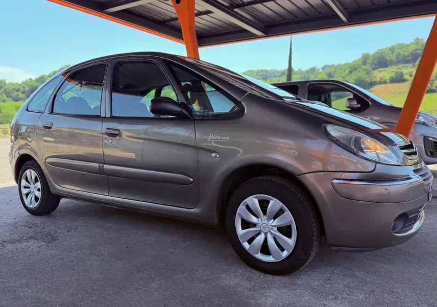Vue 3/4 avant droit d'une Citroën Xsara Picasso beige stationnée sous abri, avec jantes argentées et logo Picasso visible sur la porte.
