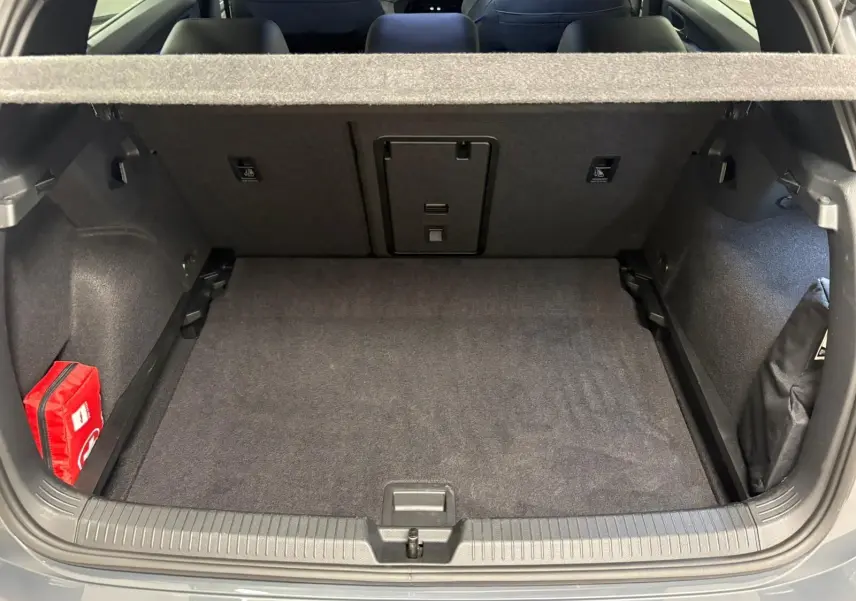 Coffre ouvert vu de l'arrière d'une Volkswagen Golf 1.5 eTSI gris dauphin, avec tapis noir et trousse de secours rouge à gauche.