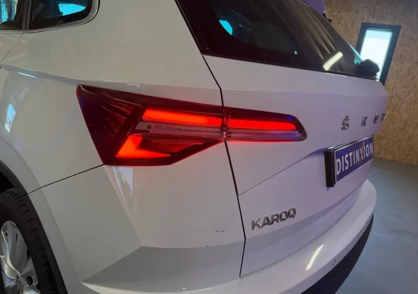 Vue 3/4 arrière droite d'un Skoda Karoq blanc 2023 avec feux arrière allumés et logo distinctif sur le hayon.