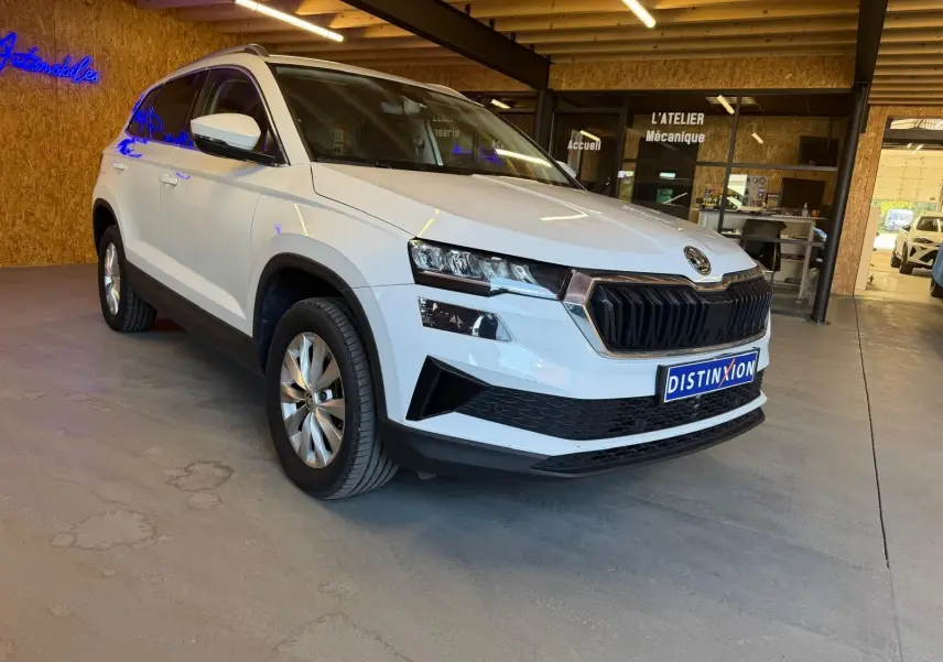 SUV Skoda Karoq blanc 2023 en 3/4 avant droit, avec calandre noire et jantes alliage visibles en intérieur showroom.
