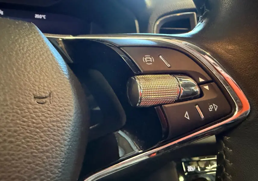 Gros plan sur les commandes au volant côté gauche du Skoda Karoq blanc 2023, avec molette métallique et boutons noirs.