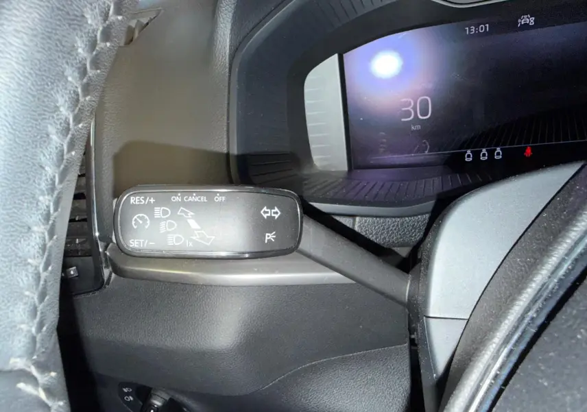 Gros plan sur la manette de régulateur de vitesse à gauche du volant du Skoda Karoq blanc, tableau de bord numérique en arrière-plan.