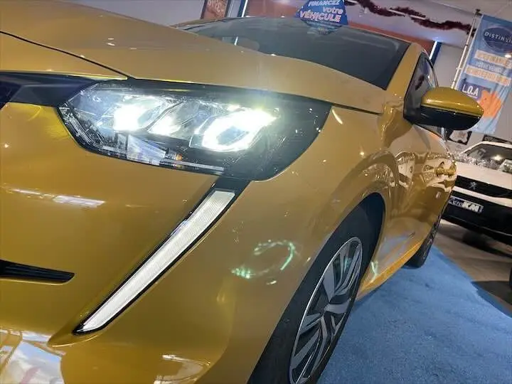 Vue 3/4 avant gauche d'une Peugeot 208 jaune Faro Metal avec phares LED allumés en intérieur showroom.