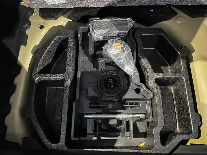 Vue en plongée sur le compartiment de rangement sous le coffre d'une Peugeot 208 jaune Faro Metal, montrant les outils d'origine.