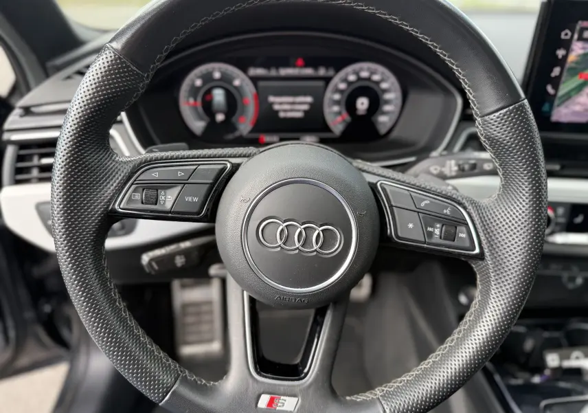 Volant en cuir perforé avec logo Audi et badge S-Line, tableau de bord numérique en arrière-plan, intérieur d'Audi A4 Avant 2023.